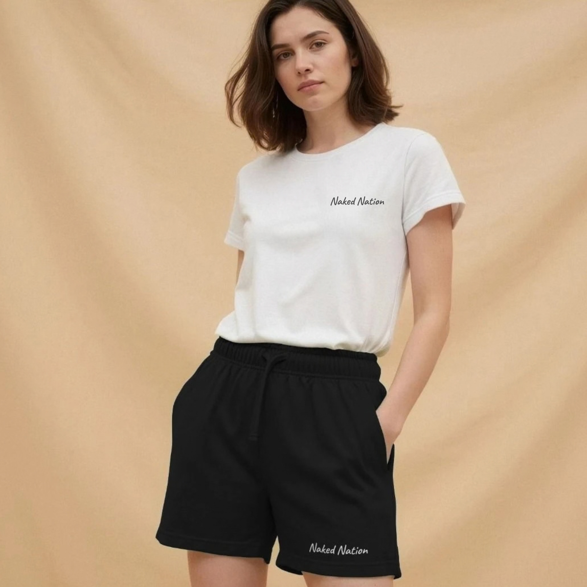 A woman modeling a white crewneck t-shirt and black drawstring shorts, both featuring the 'Naked Nation' logo printed in white script. She is standing against a warm beige fabric backdrop.