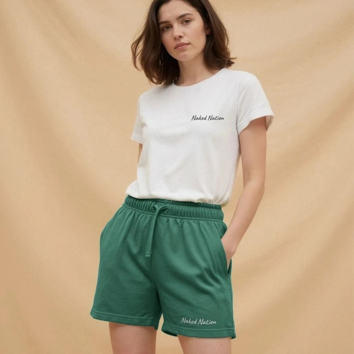 A woman modeling a white crewneck t-shirt and green drawstring shorts, both featuring the ' Naked Nation' logo, set against a warm beige backdrop.